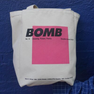 BOMB archival tote bag featuring a reproduction of a pink square. Tote bag is pictured against a blue brick wall.