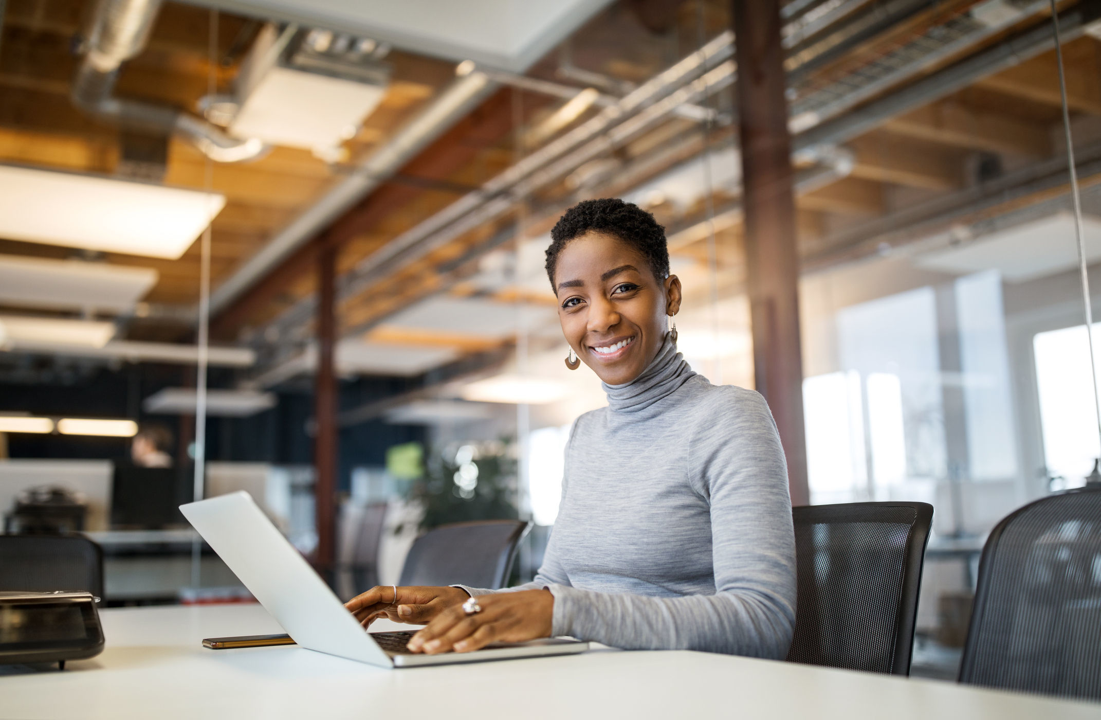 A woman looks at the camera and smiles. Before her is her laptop computer. Behind her is a modern open workspace.
