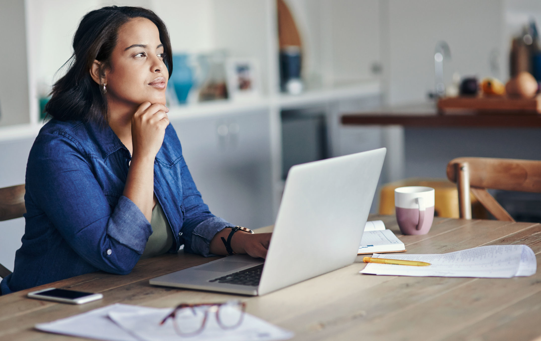 A young woman seated at a table in her kitchen thinks about something while using a laptop computer.