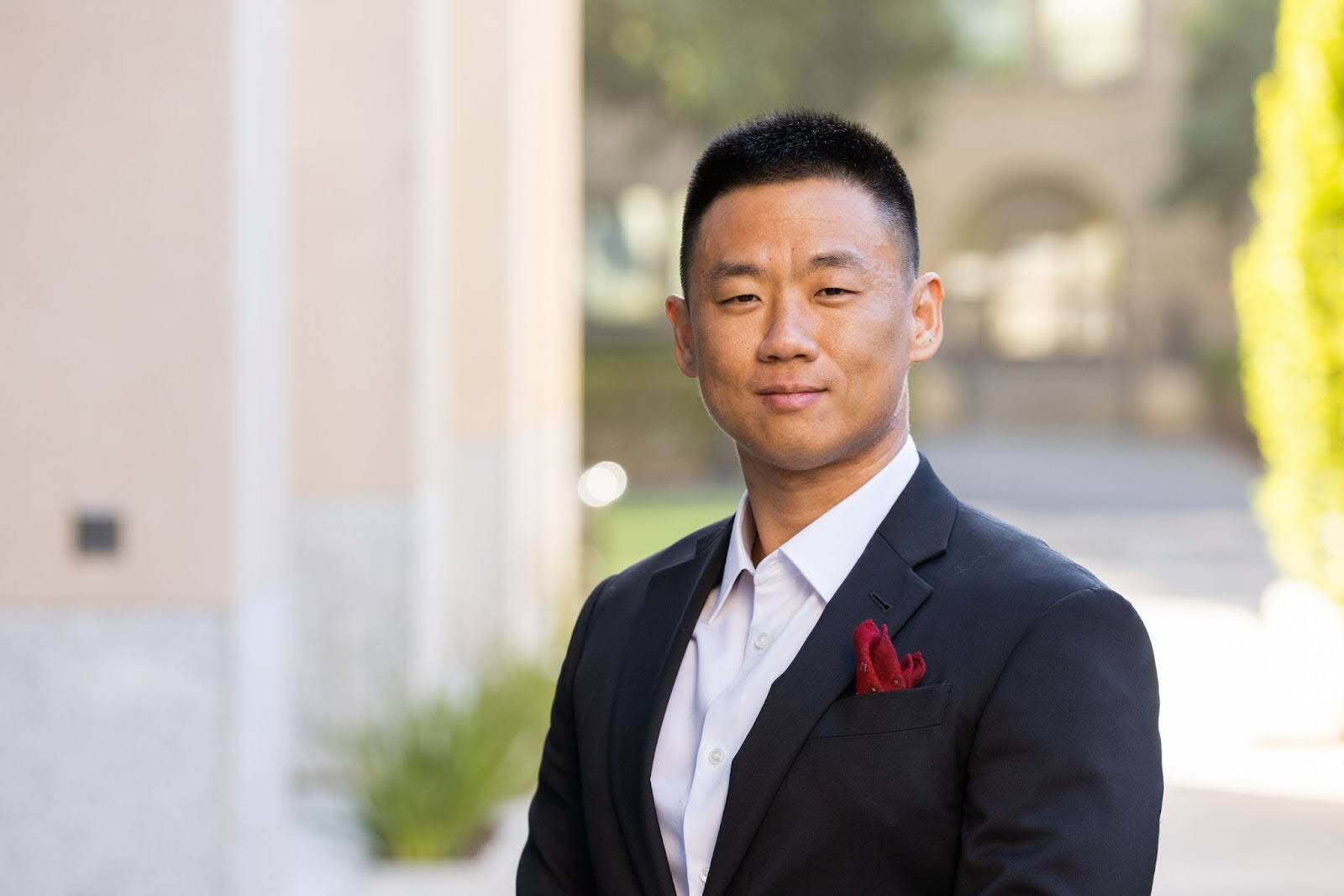 Haotian Bai, the CEO and Founder of Patriot Conceptions, seen from the waist up. He is an Asian man with short dark hair and he is wearing a white collared shirt and a black suit jacket with a red handkerchief in the breast pocket.