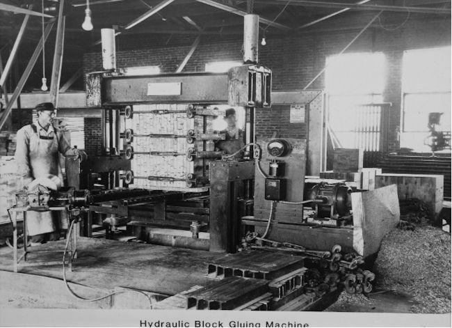 A historic photo from John Boos & Co. of its hydraulic block gluing machine with two production workers pictured next to it.
