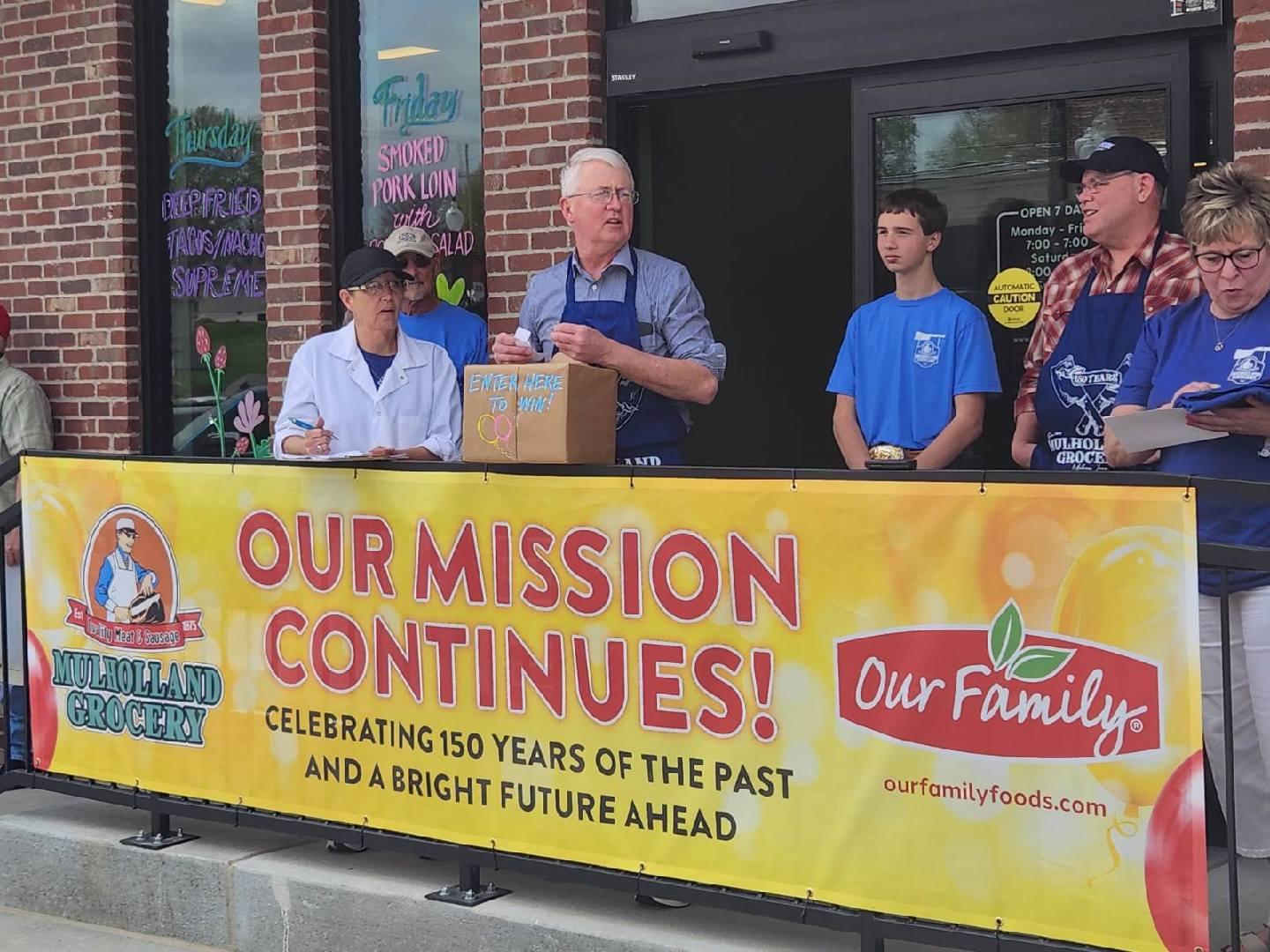 A group of people, including grocery store owner Tom Mulholland, standing behind a banner. The banner is large and yellow, reading "OUR MISSION CONTINUES! Celebrating 150 Years of the Past and a Bright Future Ahead" between the logos for Mulholland Grocery and Our Family Foods.