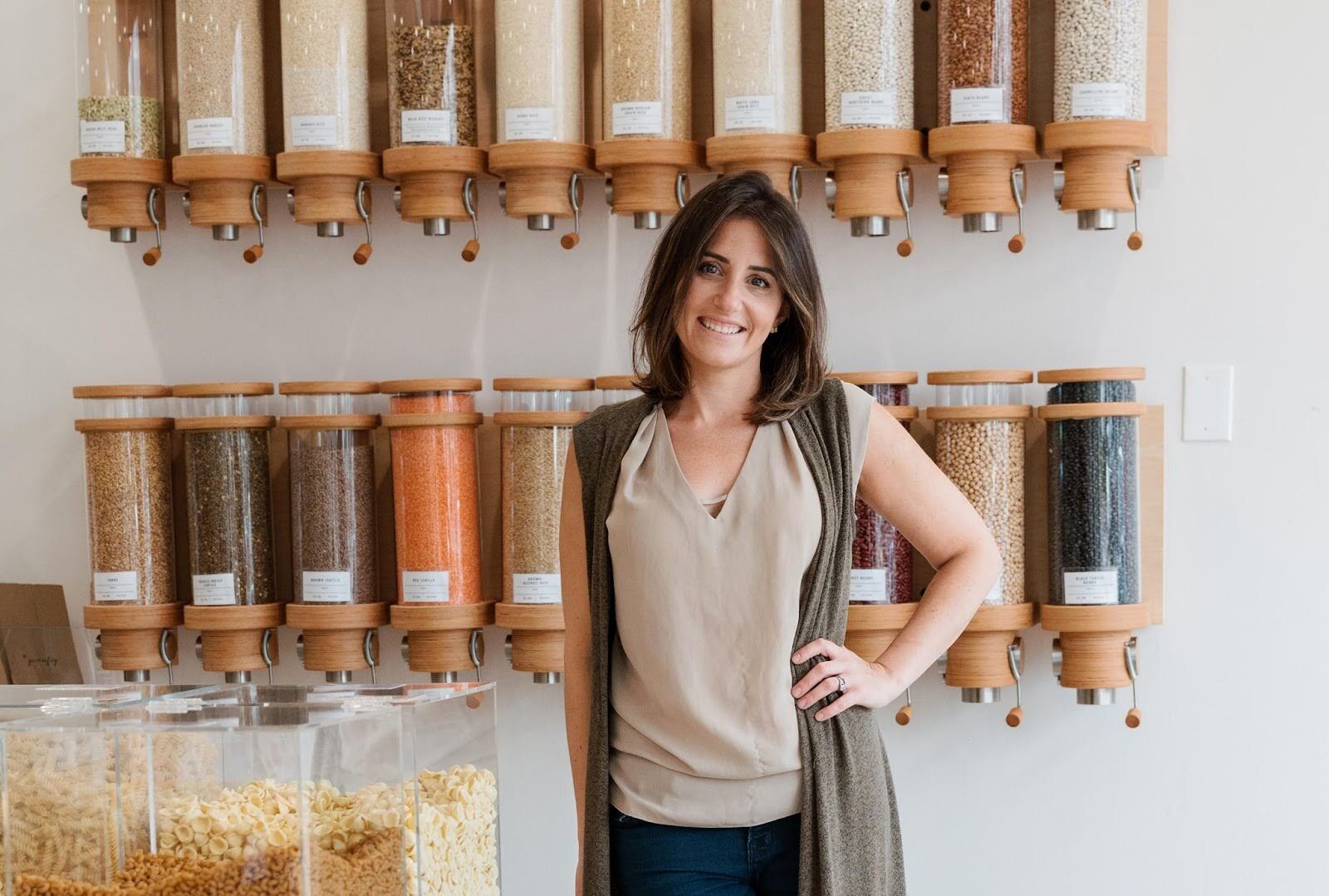 Rachel Garcia, the Founder of Dry Goods Refillery, stands in front of a wall of dispenseries of grain. Rachel stands with one hand on her hip and a smile on her face. She has shoulder-length brown hair and brown eyes, and she's wearing a pale beige V-neck T-shirt under a gray sleeveless cardigan. Clear containers of cereal are lined up to the left of her.