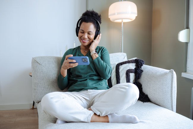 A woman sits on a couch in her home. She is wearing headphones and watching a video on her phone.