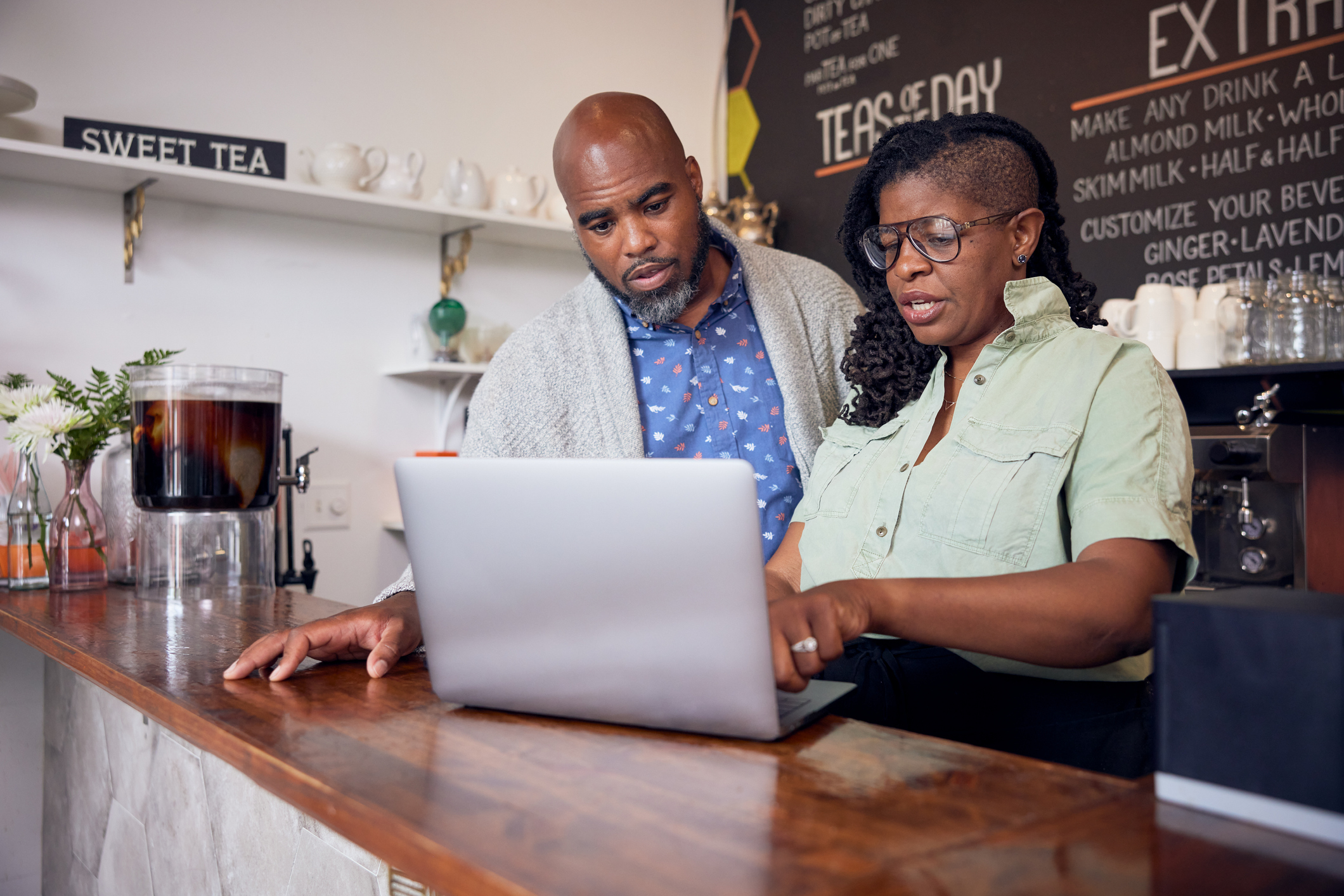 A man and a woman stand at a wood-topped counter in a cafe and look at the screen of an open laptop. The woman stands on the right and has her mouth open as if she's saying something. Her hands are on the keyboard and she wears a pale green button-up shirt and glasses. The man leans forward slightly to look at the laptop; he is bald with a graying beard and he wears a blue-and-white polka dot shirt and a light gray jacket. Behind the couple are a coffee machine, shelves holding mugs and glasses, and a large blackboard with a menu listing teas of the day and various milk additions.