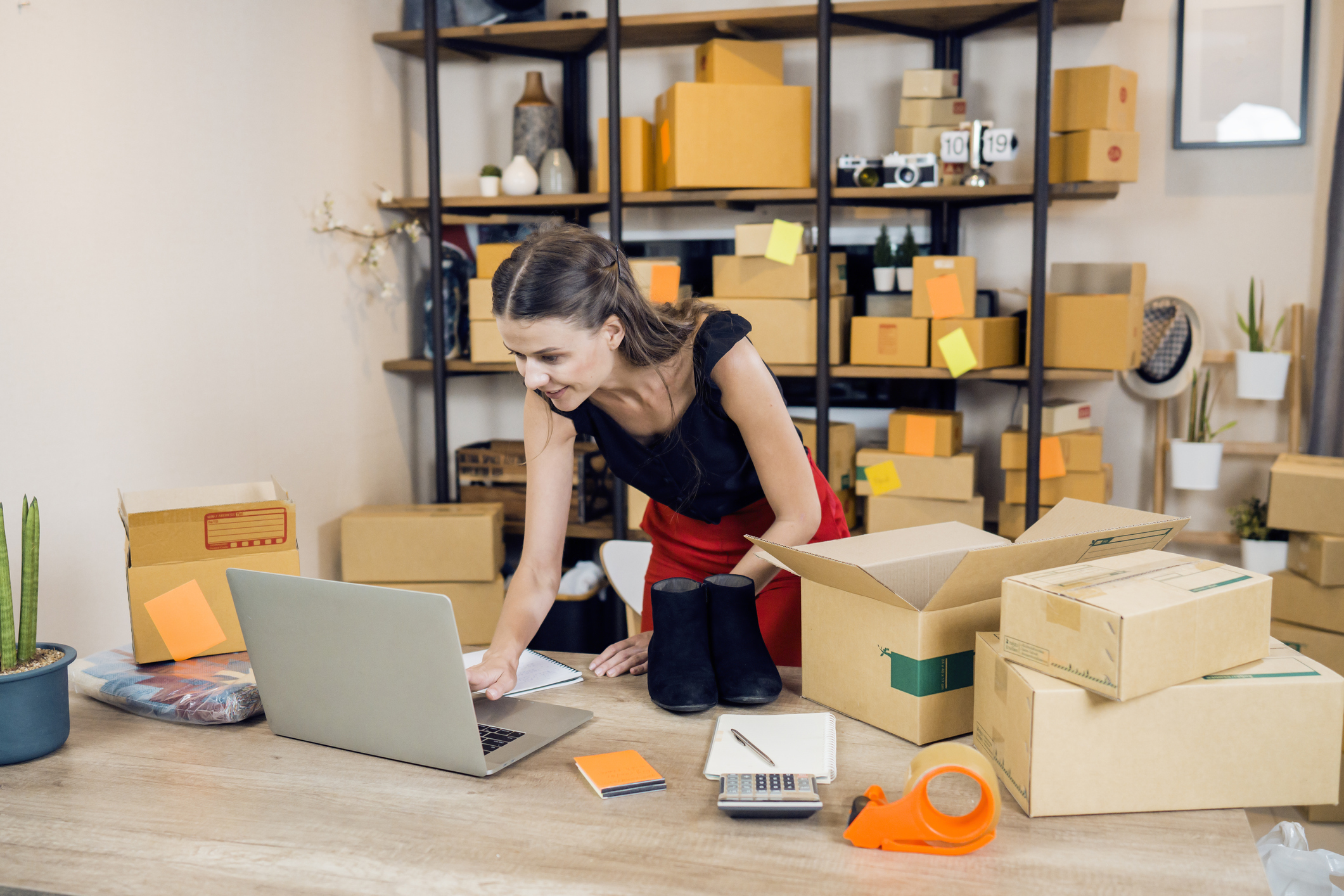 A woman leans over an open laptop on a wooden table. The woman has long dark hair and wears a black shirt and red pants. She has one hand on the laptop's trackpad. Cardboard boxes sit on either side of the laptop on the table and fill the shelving unit behind the woman. Also on the table are a calculator, a roll of packing tape in an orange holder, a notebook and pen, and a pair of black boots ready to be packed into the open box next to it.