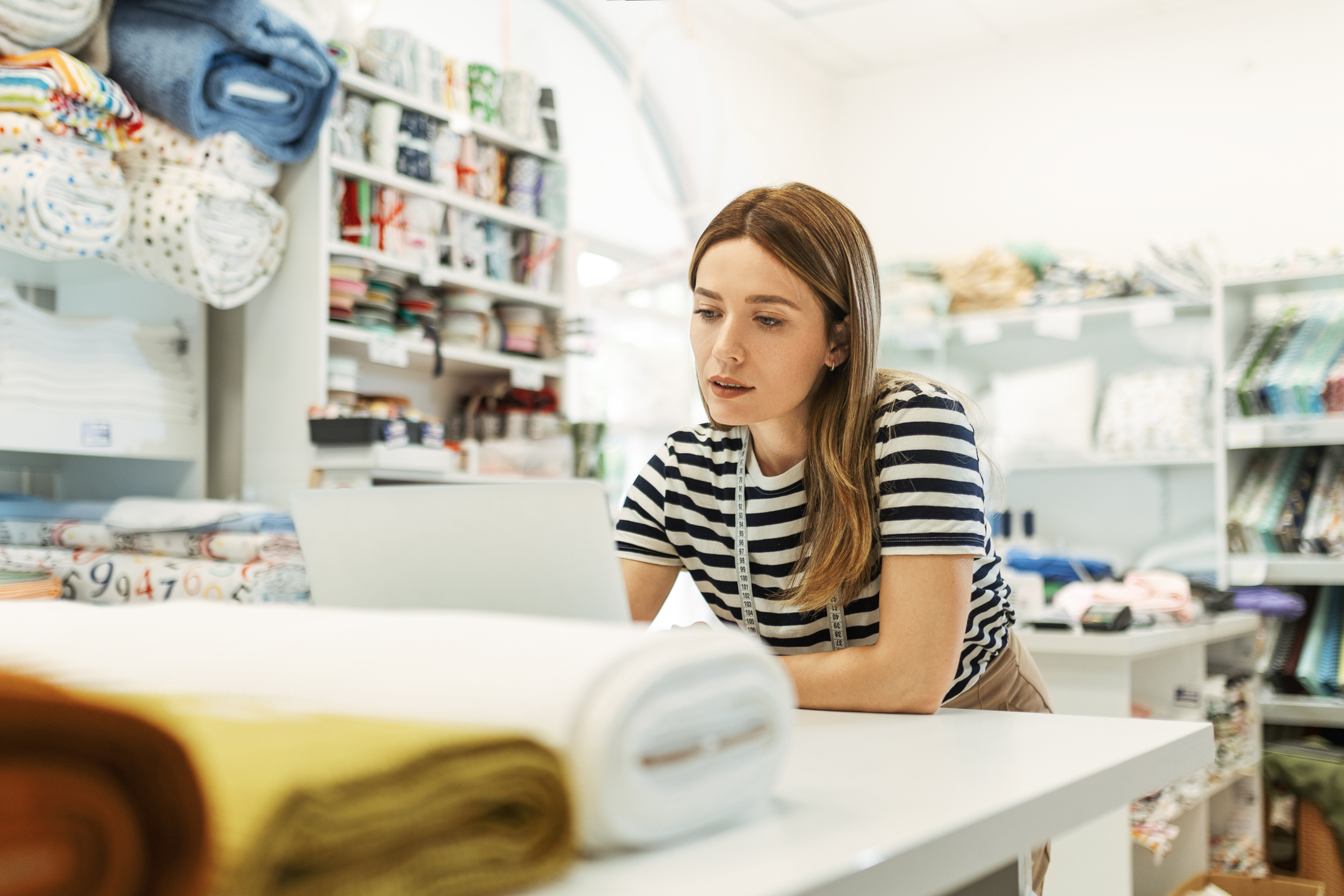 A young woman sits at a table in her workshop and works on a laptop. She has center-parted honey blonde hair and wears a black-and-white striped shirt and a measuring tape draped around her neck. The workshop has white walls and is filled with shelves and cubbies of rolls of fabric and thread. In the foreground are white, yellow, and dark orange fabric, sitting in rolls on the table.