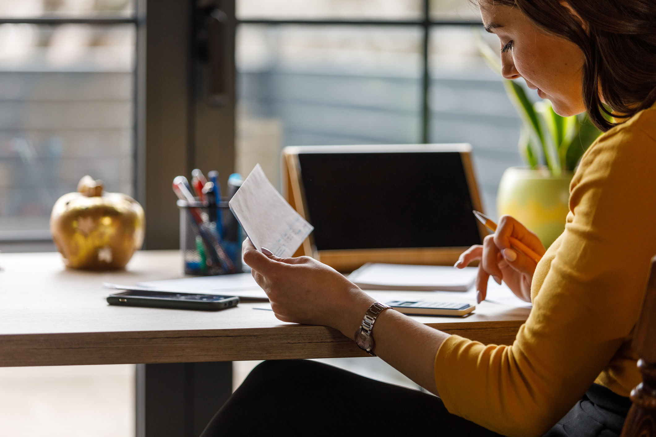A young woman is seated at a table. She is holding up a piece of paper in her right hand and using a calculator in her right hand to help her determine how much she owes to the IRS.