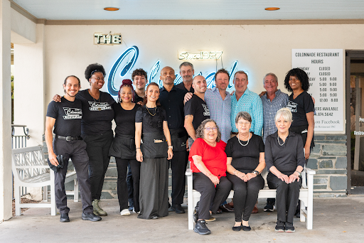 The team of employees at The Colonnade restaurant standing and sitting together, smiling for the camera. Behind them, partially obscured, it a large neon side reading "The Colonnade," with "The" in gold" and "Colonnade" in large blue cursive.