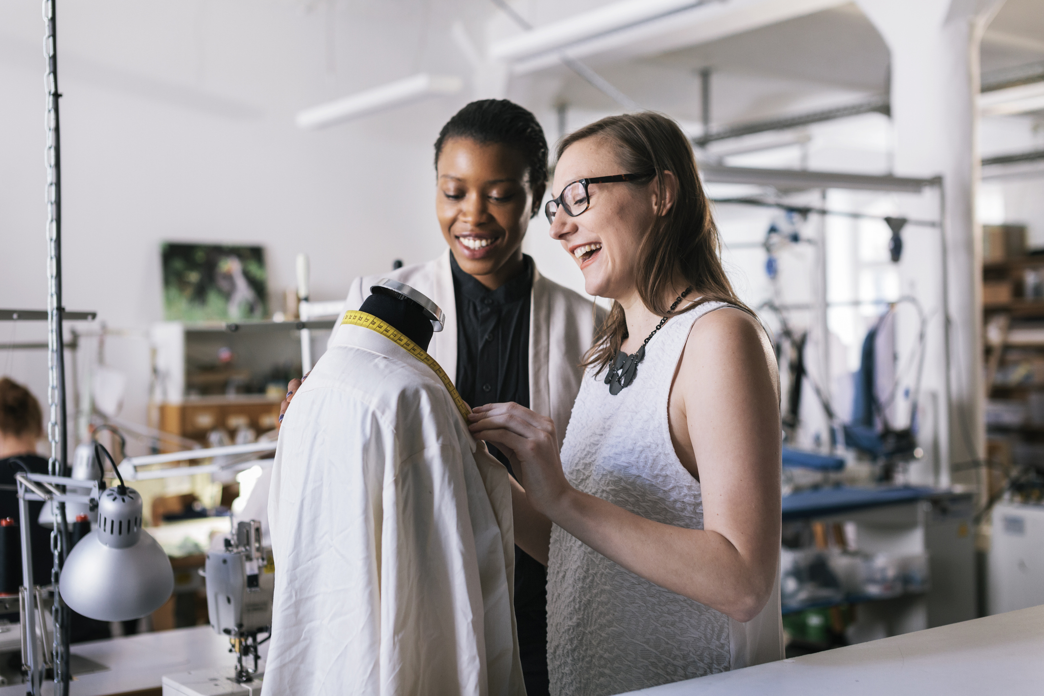 Two women stand smiling in front of a headless mannequin draped with a loose-fitting white blouse. The woman on the left has short dark hair and wears a cream-colored jacket over a black button-up shirt. The woman on the right has long dark blonde hair and wears glasses and a white textured tank top. The woman on the right is examining a measuring tape that she has draped around the back of the mannequin's neck. In the background, out of focus, is a large studio space containing a few sewing machine stations and shelving units.