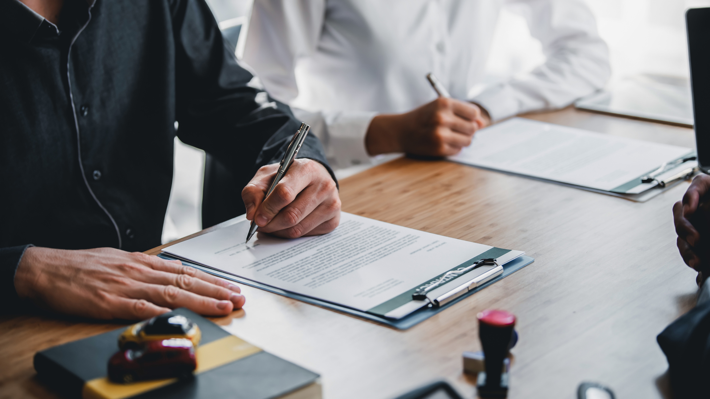 A below-the-neck shot of two people in button-up shirts sitting side by side at a table signing paperwork. The paperwork is attached to two clipboards. Also on the table but out-of-focus are a book and a rubber stamp.