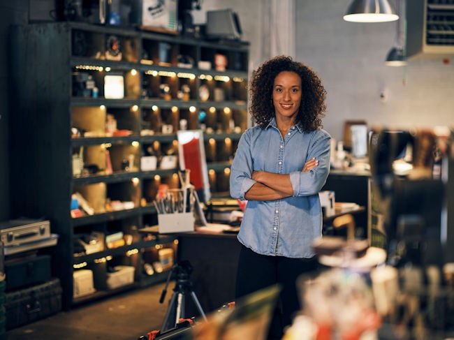 A woman smiles at the viewer. She is standing in the setting of her studio.