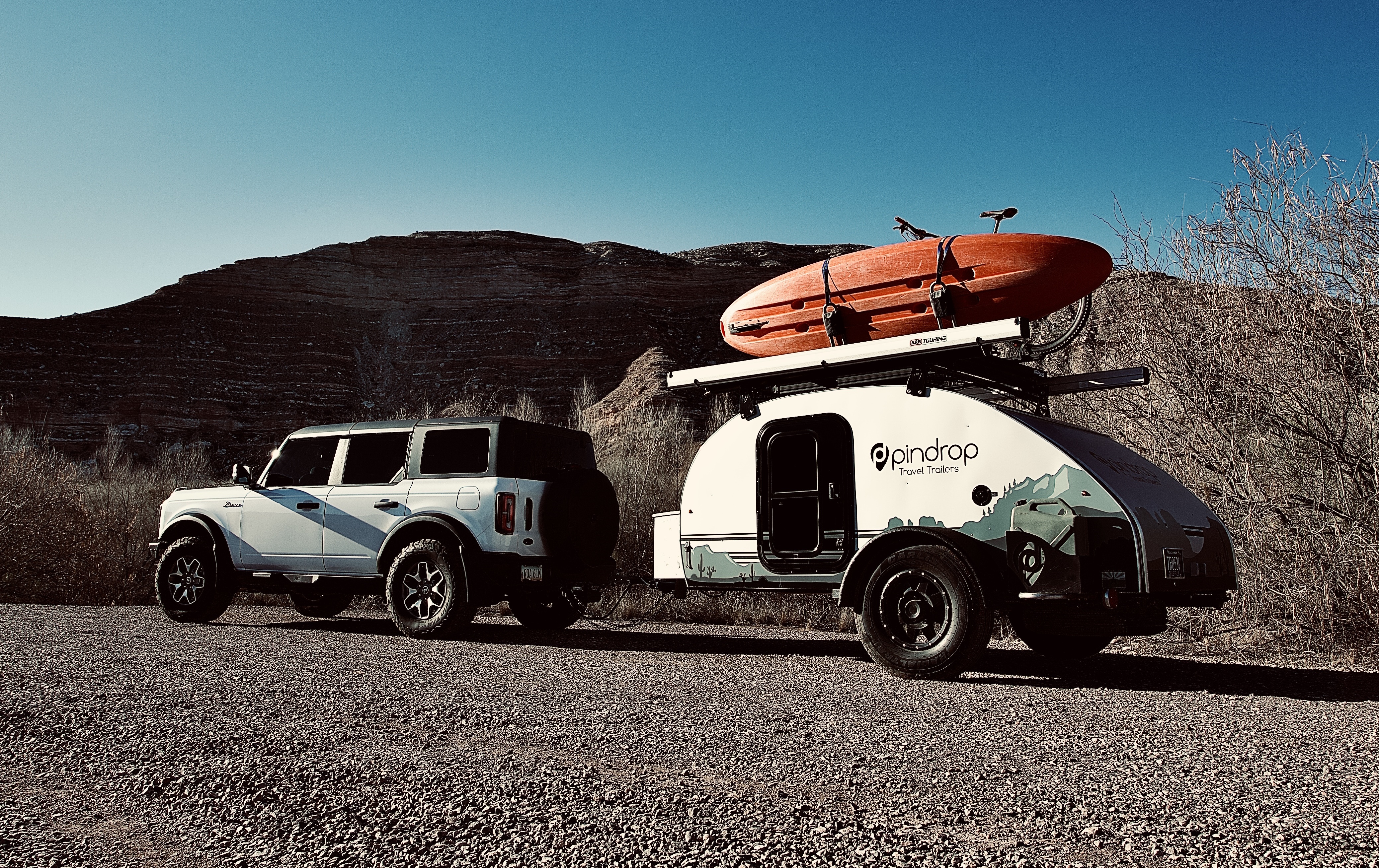A wide shot of a Pin Drop micro camper trailer tethered to the back of a gray truck. The micro camper is shaped like a half-circle laying on its side, with two wheels positioned near the rear and an orange kayak mounted on top. The camper is painted with a desert landscape in shades of green, and the Pin Drop logo appears next to the camper's door. The camper and truck are parked in a desert next to scrub brush, with a mountain looming in the distance.