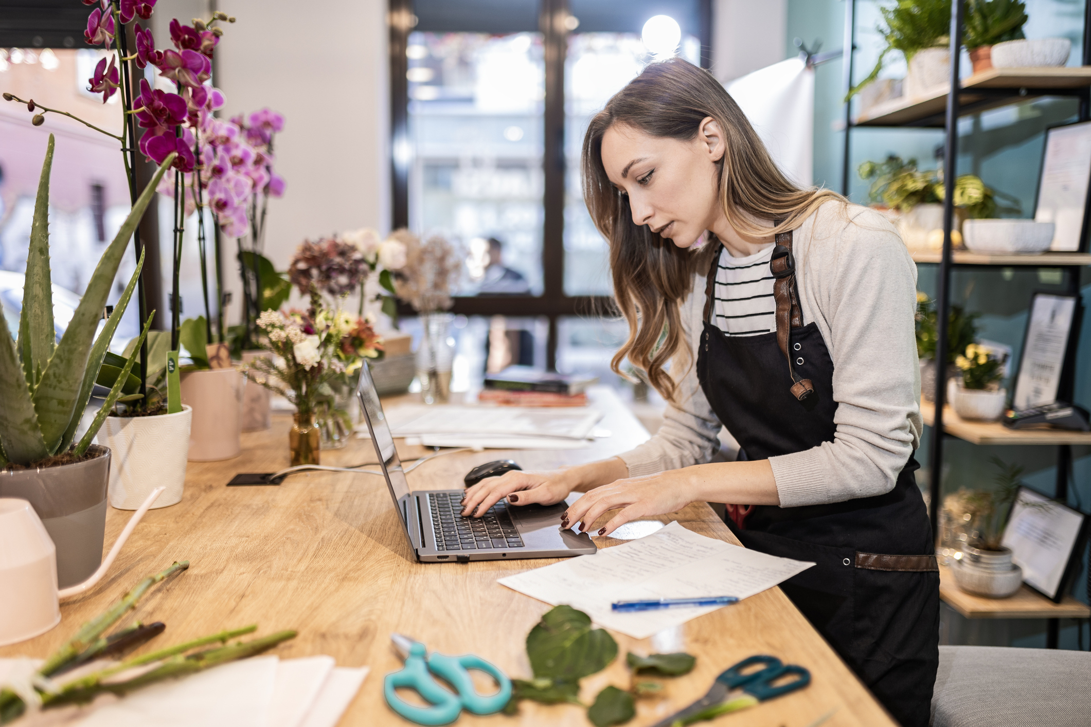 A woman stands at a table in a workshop and works on a laptop. The woman has long brown hair and wears a white long-sleeved shirt and a black apron. Elswhere on the workshop table are pruning shears and potted plants.