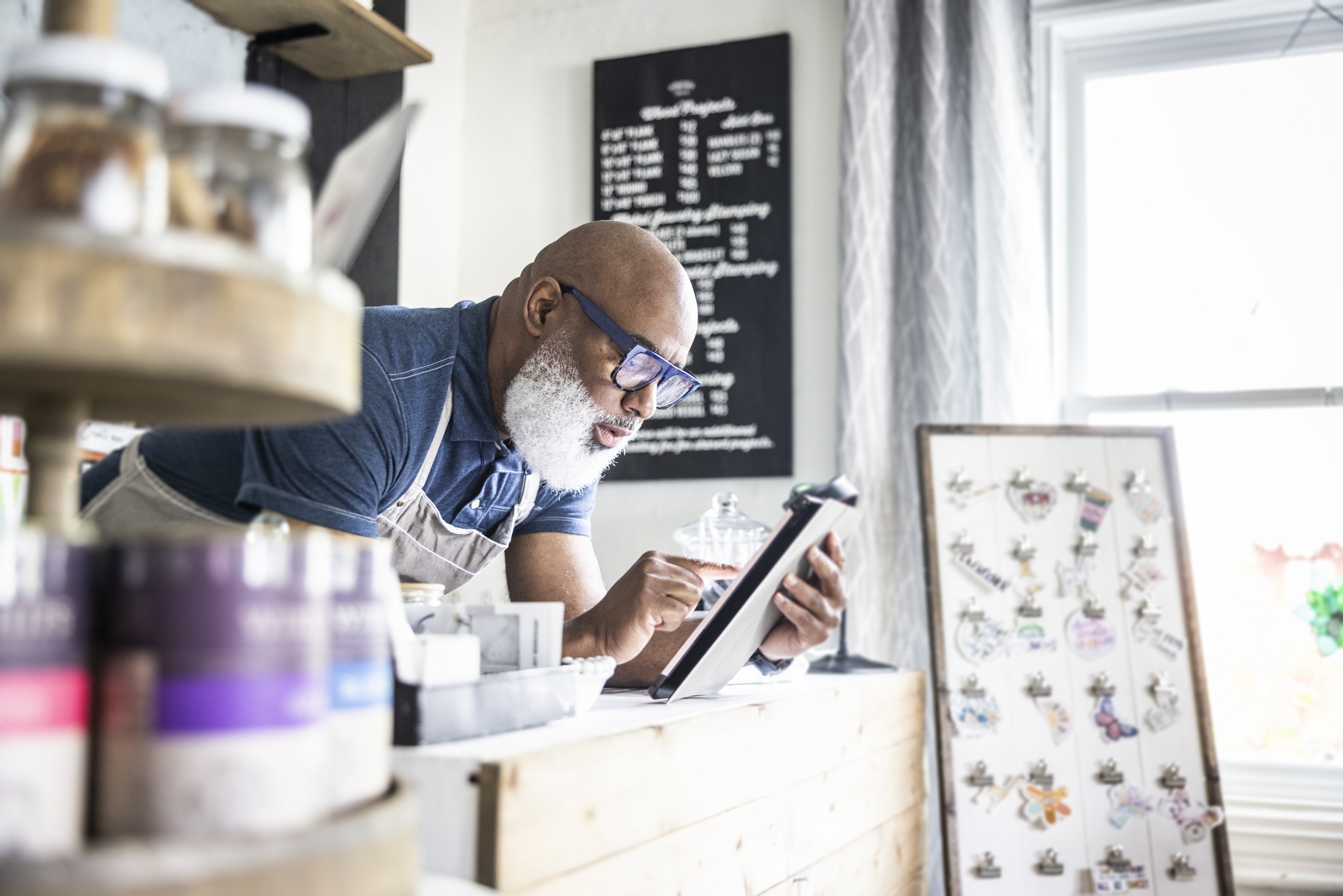 A business owner with a beard and glasses looking at a tablet inside his cafe.