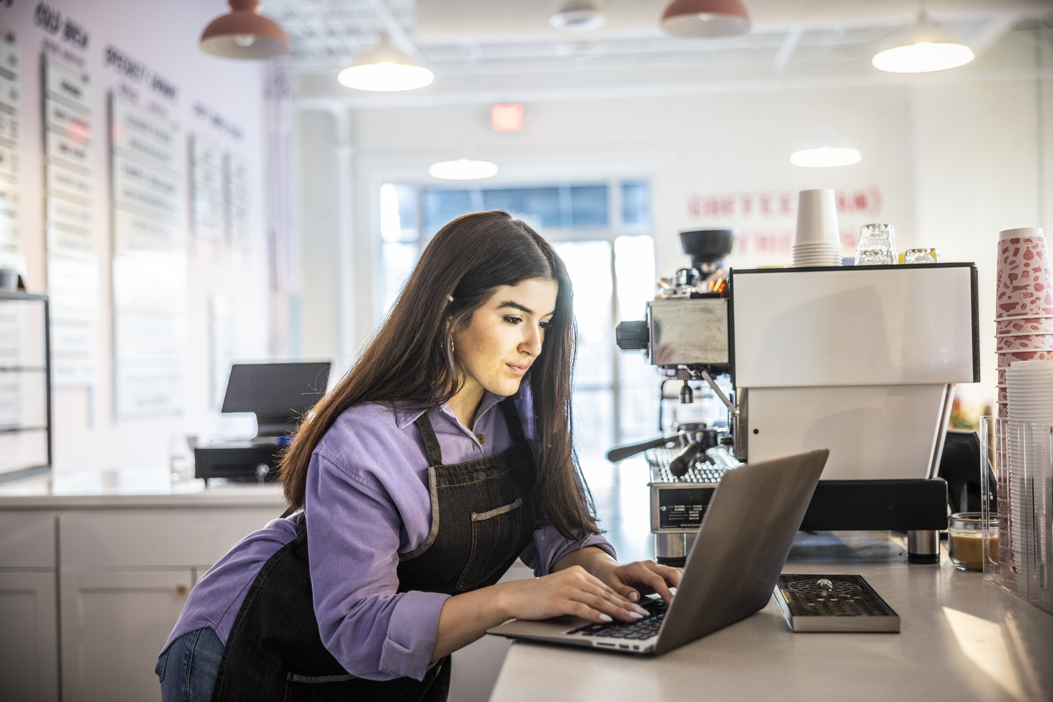 A woman stands behind the counter at a cafe, typing on a laptop. The woman has long brown hair and she wears a dark denim apron over a lavender button-up shirt and jeans. The cafe is all white, with the shop's offerings listed on signs along one of the walls. The L-shaped counter holds a large coffee machines and stacks of paper cups.