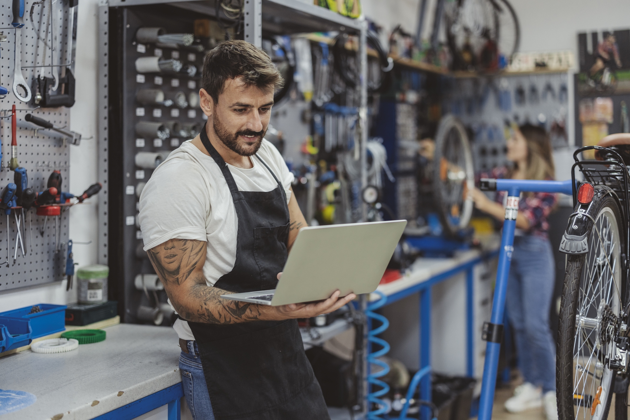 A man in an apron stands in a bicycle repair shop, holding a laptop. The man has dark hair and a beard, and he has sleeve tattoos covering both arms.