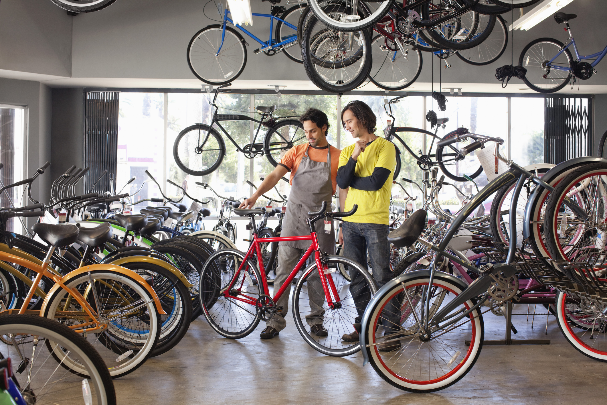 Two men stand in a large showroom filled with bicycles of various colors. The man on the left, who wears an orange shirt and gray apron, uses his hands to hold up a red bike. The man on the right, who wears a yellow T-shirt over a long-sleeved navy blue shirt, looks at the red bike with interests. Several more bikes hang from the ceiling of the showroom.