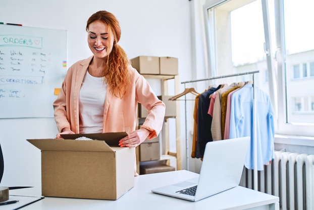 A woman closes a box she was packing. In the background are more boxes, a whiteboard, and a rack of clothing.