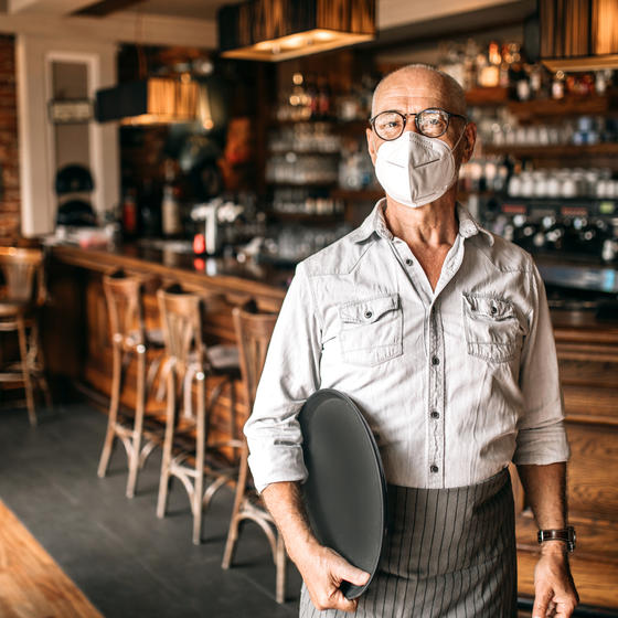 A man wearing a face mask stands in a restaurant and holds a serving platter. In the background is a well-stocked bar.