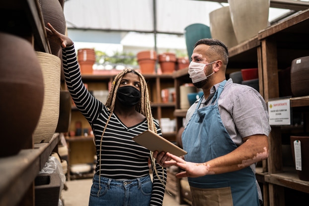 A Black woman gestures to a shelf of planters as she talks with an apron-wearing man with vitiligo. Both people are wearing face masks.
