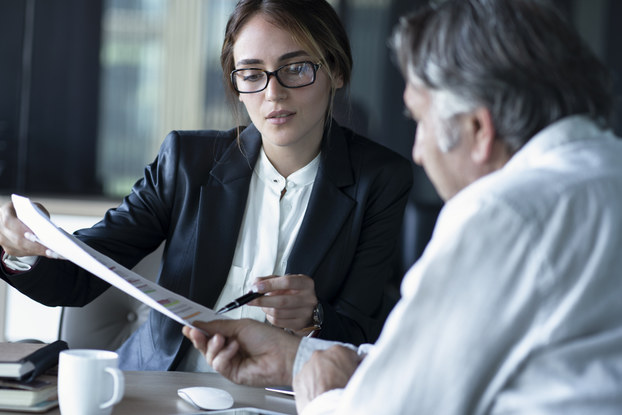 A woman wearing glasses and a blazer shows a paper to an older man in a collared shirt. The woman uses a pen to point to something written on the page.