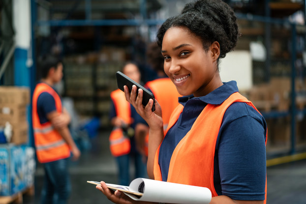 A woman in a dark blue polo shirt and neon orange vest holds her smart phone near her face with one hand and holds a clipboard with the other hand. In the background, employees dressed similarly to the woman are standing around a warehouse filled with cardboard boxes and packages of bottled water.
