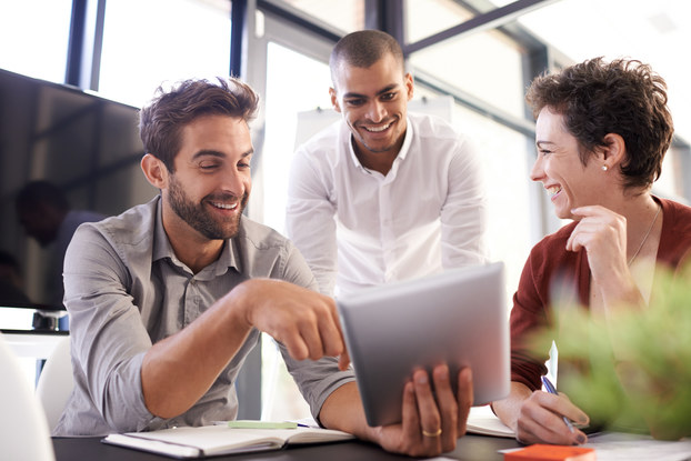 Three people (two men and a woman) surround a table. One of the men holds up a tablet and shows something to his two colleagues.