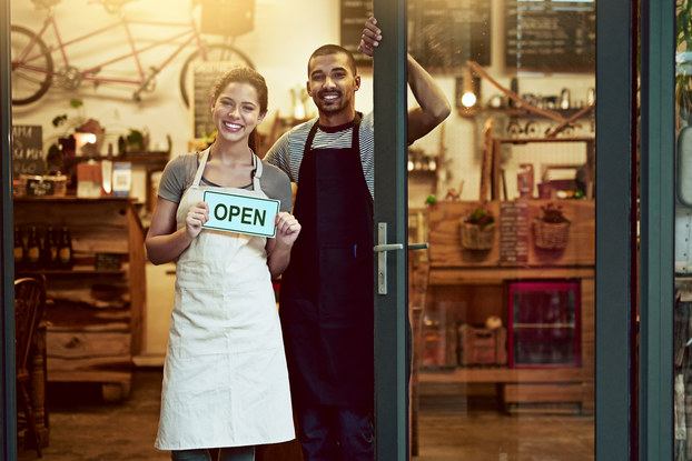 A man and a woman stand smiling in the doorway of a shop. The woman holds an OPEN sign.