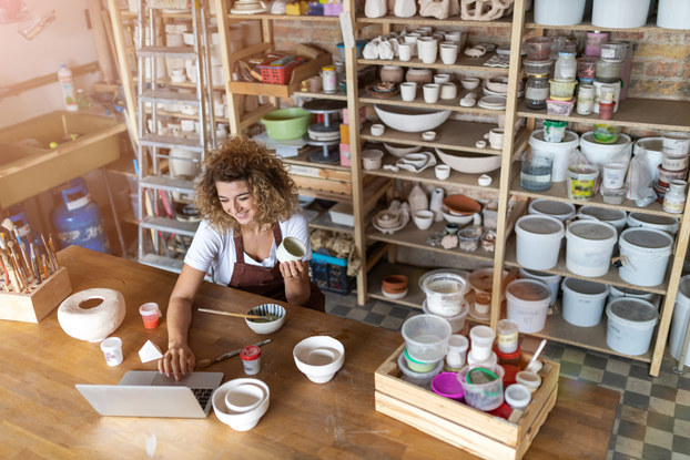 A woman works on her laptop at a large wooden table scattered with pottery. Behind her are shelves of handmade bowls, pots, buckets, and other pieces of pottery.