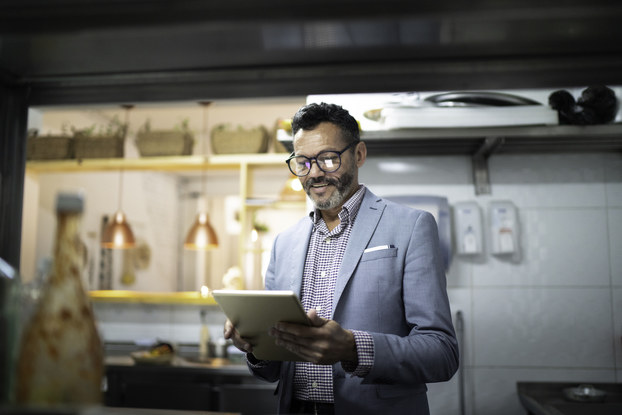 A man stands in a restaurant kitchen and consults a tablet.