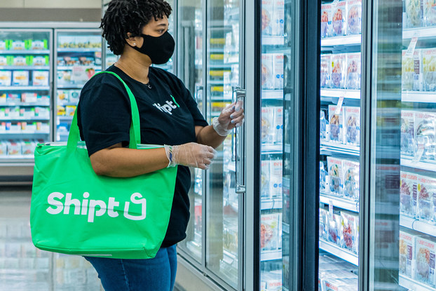 A Shipt employee wearing a mask looks through the frozen food section of a partnering grocery store for requested food items, which will be placed in the large green bag hanging from the shopper's shoulder.