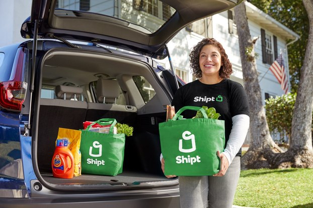 A Shipt driver unloads groceries in green Shipt bags from the trunk of a blue car.