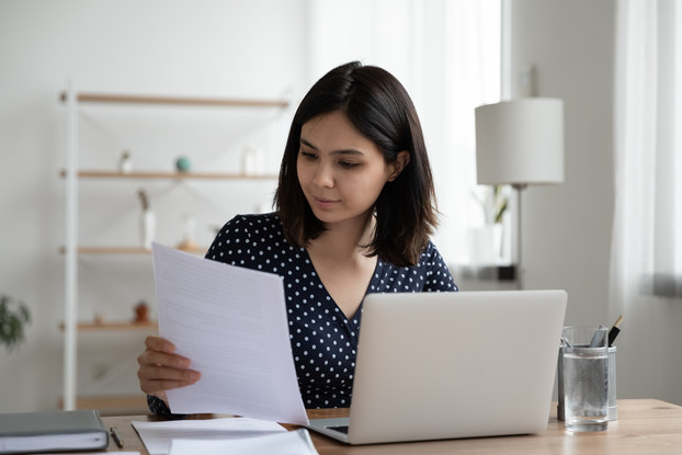 A young woman considers the contents of a piece of paper as she sits in front of a laptop.