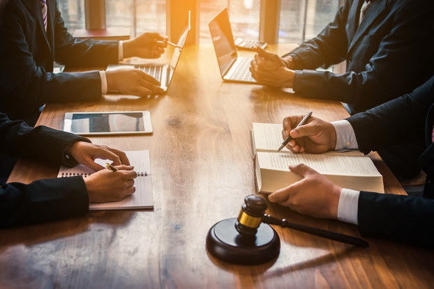 Four people, seen only from the shoulders down, sit around a wooden table. Two of the people have their hands on laptops; the other two people have their hands on open books. In the foreground sits a judge's gavel.