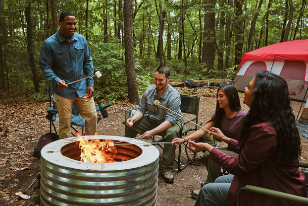 A group of friends roasts marshmallows around a metal firepit. They're sitting in foldable chairs in a forest clearing. A large tent is set up in the background.