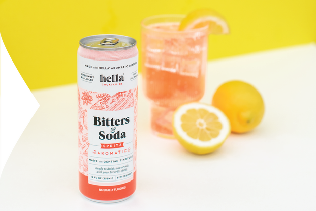 Can of Hella Cocktail Co. bitters & soda on a table with a full glass and a lemon,