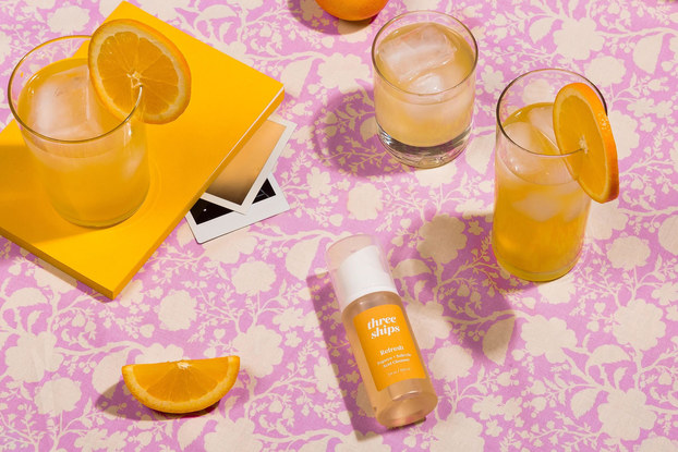 A bottle of Three Ships Refresh cleanser lays on its side next to a cocktail and pieces of fruit.