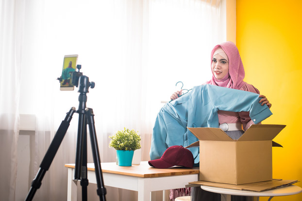 A woman shows off a light blue jacket on a video that she films on a smartphone attached to a tripod.