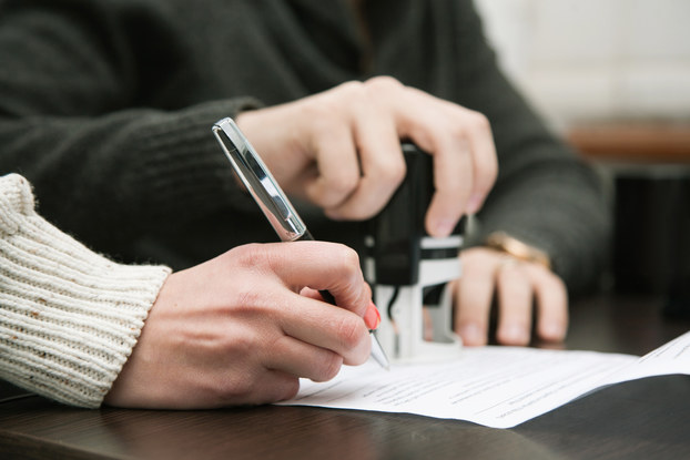 Close-up on two sets of hands. One person's hand signs a contract while the other person readies a stamp for the form.