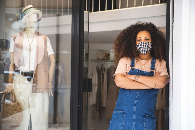 woman business owner wearing mask standing in doorway of her retail shop