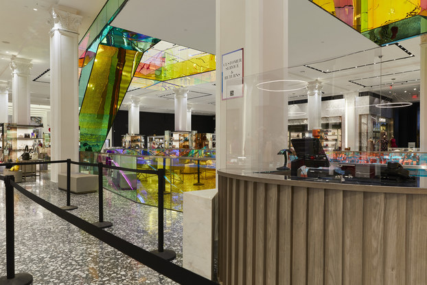 A client services desk behind a safety glass. Black ropes and poles outline the customer queue. In the background are displays with purses and the underside of the mirrored escalator.