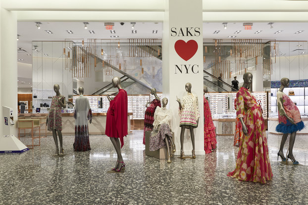 Nine mannequins model bright summer dresses. Several of the mannequins are clustered around a column bearing the message "SAKS HEARTS NYC."