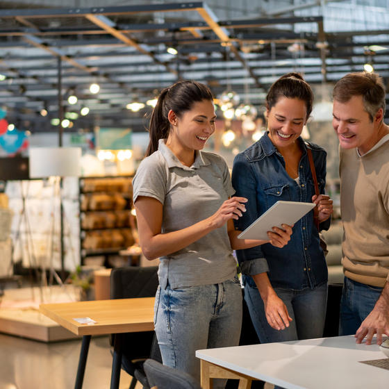 Three smiling people stand in a hardware/home improvement store. The woman on the left holds a tablet, which the man and woman on the right examine..