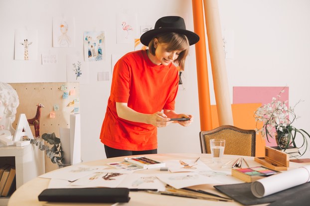 A woman in a bright orange shirt and black brimmed hat uses her smartphone to take a picture of art scattered on the table in front of her.