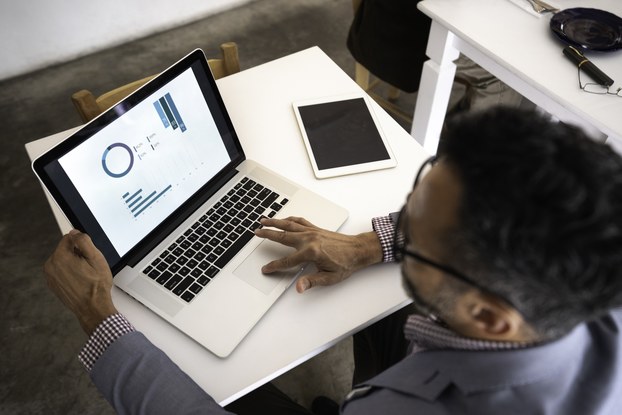 A man, seen from above, looks at a series of charts on a laptop screen. On the table next to his laptop is a smart tablet.