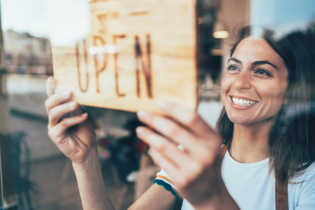 Close-up of a young woman turning a sign in a window to read OPEN. Unlike most OPEN signs, this one is wooden and the words are burned into the wood.