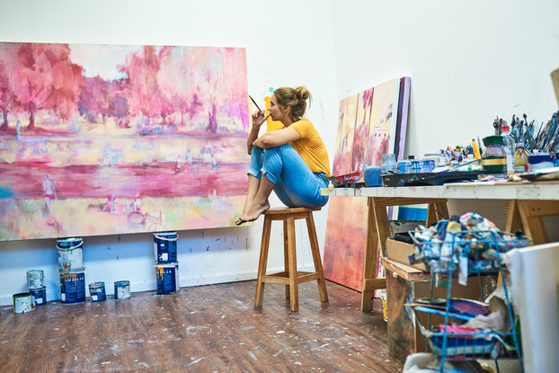 Woman artist sitting in her studio holding a paintbrush and looking at her painting.