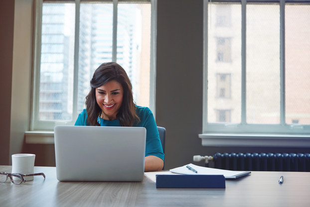 A woman sits at a large table and reads something on her laptop with a smile.