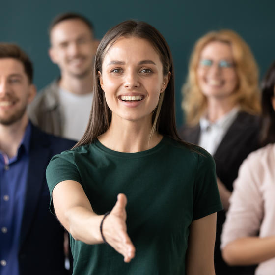 A diverse group of people stand facing the viewer. In the front of the group, a woman extends her hand for a handshake.
